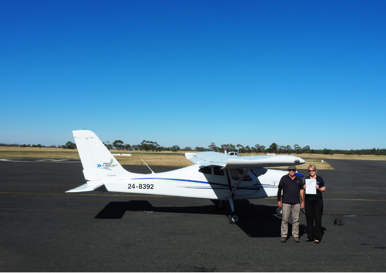 Two people standing in front of a small airplane on a runway with a clear blue sky.